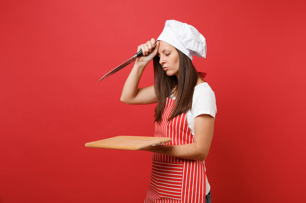 Νοικοκυρά θηλυκό σεφ μάγειρας ή φούρναρης με ριγέ ποδιά, λευκό t-shirt, Τόκα σεφ καπέλο απομονώνονται σε φόντο κόκκινο τοίχο. Οικονόμος γυναίκα εκμετάλλευση ξύλο κοπής, μαχαίρι. Χλευάσουμε επάνω το αντίγραφο διαστημικής έννοιας - Φωτογραφία, εικόνα
