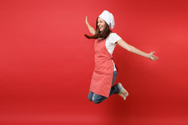 Housewife female chef cook or baker in striped apron white t-shirt, toque chefs hat isolated on red wall background. Full length portrait housekeeper woman jumping high up. Mock up copy space concept - Valokuva, kuva