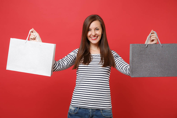 Portret van lachende jonge vrouw in gestreepte kleding bedrijf pakketten zakken met aankopen na winkelen geïsoleerd op rode muur achtergrond. Mensen oprechte emoties, lifestyle concept. Bespotten kopie databaseruimte - Foto, afbeelding