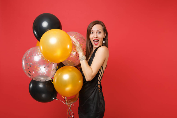 Aufgeregtes junges Mädchen in kleinem schwarzen Kleid feiert mit Luftballons auf rotem Hintergrund. St. Valentin, internationaler Frauentag, frohes neues Jahr, Geburtstagsparty-Konzept - Foto, Bild