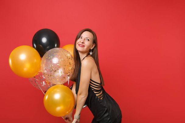 Усміхаючись молоду жінку в Маленька чорна сукня святкування дивлячись і тримають повітряних кулях ізольовані на яскраво-червоним тлом. До дня Святого Валентина день, з новим роком, день народження макет holiday party концепції - Фото, зображення