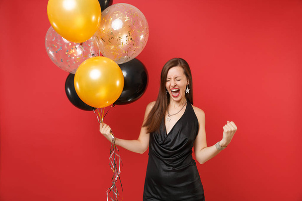 Joyful young girl in little black dress celebrate clenching fist like winner holding air balloons isolated on red background. St. Valentine's Day, Happy New Year birthday mockup holiday party concept - Foto, Imagen