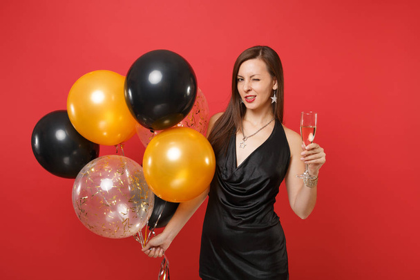 Passionate woman in black dress celebrating blinking holding glass of champagne air balloons isolated on red background. International Women's Day Happy New Year birthday mockup holiday party concept - 写真・画像
