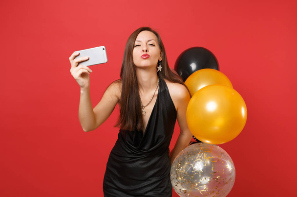 hübsche Frau in schwarzem Kleid weht Lippen, Luftkuss halten Luftballons machen Selfie-Foto auf Handy isoliert auf rotem Hintergrund. Frohes neues Jahr Geburtstag Attrappe Urlaub Party-Konzept - Foto, Bild