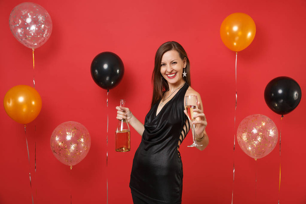 Laughing young girl in black dress celebrating holding glass, bottle of champagne on bright red background air balloons. International Women's Day Happy New Year birthday mockup holiday party concept - Photo, Image