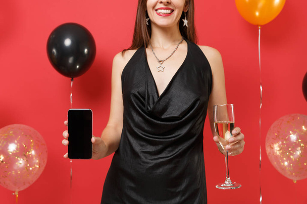 Imagen recortada de chica en vestido negro sosteniendo copa de champán, teléfono móvil con pantalla vacía en blanco negro sobre globos de aire de fondo rojo brillante. Feliz Año Nuevo cumpleaños mockup concepto de fiesta de vacaciones
 - Foto, imagen