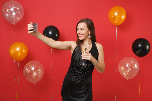 Mujer joven sonriente en vestido negro sosteniendo una copa de champán, haciendo una toma de selfie en el teléfono móvil en globos de aire de fondo rojo brillante. Feliz Año Nuevo, concepto de fiesta de cumpleaños simulada
 - Foto, Imagen
