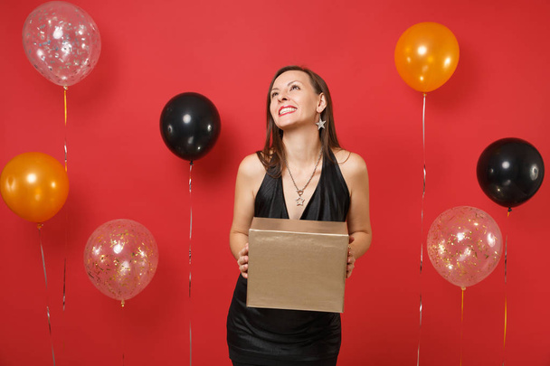 Dreamy happy girl in black dress celebrating looking up hold golden box with gift present on bright red background air balloons. Valentine's Day, Happy New Year birthday mockup holiday party concept - Foto, immagini