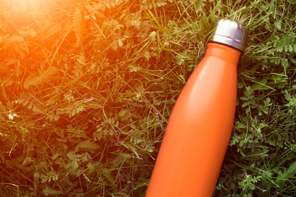 Нержавеющая термос бутылка воды, матовый оранжевый цвет. Макет изолирован на зеленом фоне травы с эффектом солнечного света
 - Фото, изображение