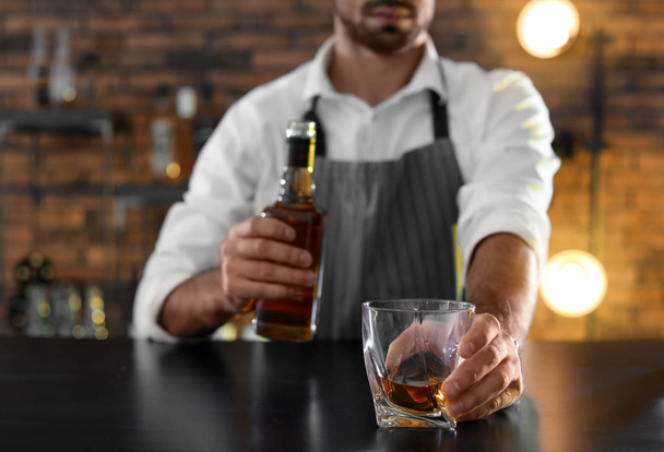 Bartender with glass and bottle of whiskey at counter in bar, closeup. Space for text - Фото, изображение