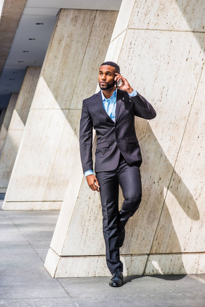 Jonge African American zakenman met baard praten op mobiele telefoon buiten het kantoor in New York City, het dragen van zwarte pak, lederen schoenen, permanent door kolom op straat, onder de zon, rondkijken - Foto, afbeelding