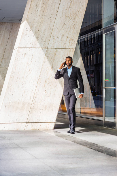 Hombre de negocios afroamericano con barba hablando por teléfono celular, viajando, trabajando en Nueva York, vistiendo traje negro, zapatos de cuero, llevando computadora portátil, saliendo del edificio de oficinas a la calle
. - Foto, Imagen