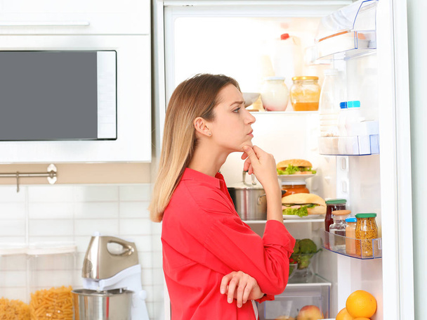 Thoughtful young woman choosing food from refrigerator in kitchen. Healthy diet - Fotó, kép