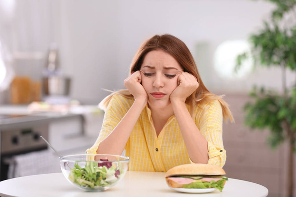 Sad young woman choosing between salad and sandwich in kitchen. Healthy diet - Foto, imagen