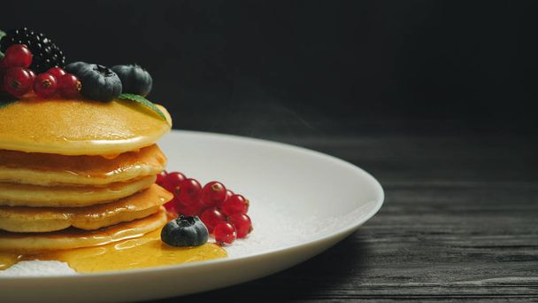 Fresh bakery. Pancakes with forest berries and honey on white plate and black smoke background. Copy space. Homemade healthy breakfast. Yummy food, dessert concept. - Foto, imagen