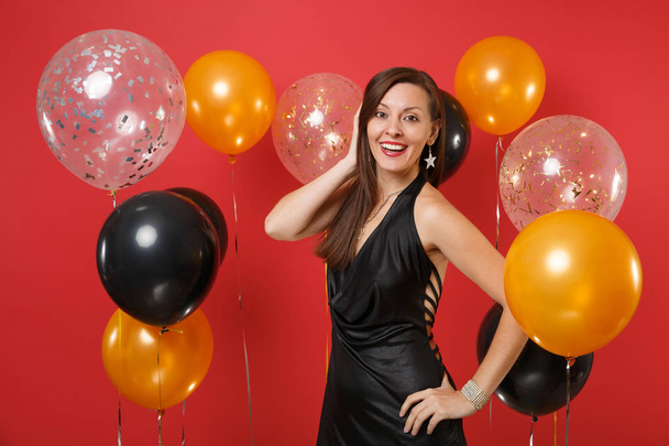 Smiling young woman in little black dress celebrating, putting hand on head on bright red background air balloons. St. Valentine's, Women's Day, Happy New Year, birthday mockup holiday party concept - Фото, изображение