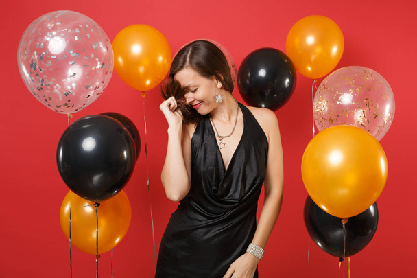 Pretty young girl with lowered head in black dress celebrating holding her hair on bright red background air balloons. International Women's Day, Happy New Year, birthday mockup holiday party concept - Fotografie, Obrázek