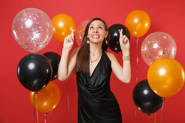 Pretty young woman in little black dress celebrating, making wish, keeping fingers crossed and looking up on bright red background air balloons. Happy New Year, birthday mockup holiday party concept - Photo, Image