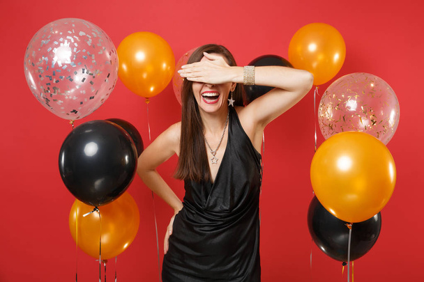 Laughing young woman in black dress celebrating covering eyes with hand on red background air balloons. St. Valentine's, International Women's Day Happy New Year birthday mockup holiday party concept - Photo, Image
