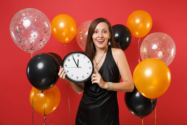 Jovem excitada em vestido preto comemorando segurar relógio redondo no balão de ar fundo vermelho brilhante. Dia Internacional da Mulher dos Namorados Feliz Ano Novo conceito festa de aniversário mockup
 - Foto, Imagem