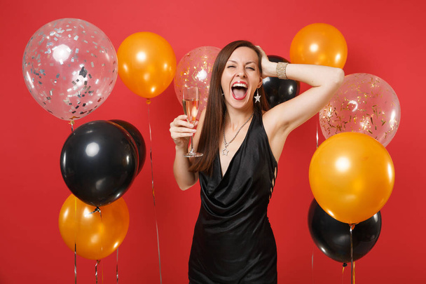 Overjoyed girl in black dress celebrating holding glass of champagne putting hand on head on bright red background air balloons. Valentine's Day, Happy New Year, birthday mockup holiday party concept - Photo, Image