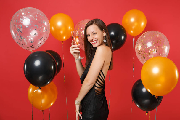 Side view of smiling pretty girl in black dress celebrating, holding glass of champagne on bright red background air balloons. Valentine's Day, Happy New Year, birthday mockup holiday party concept - Photo, Image