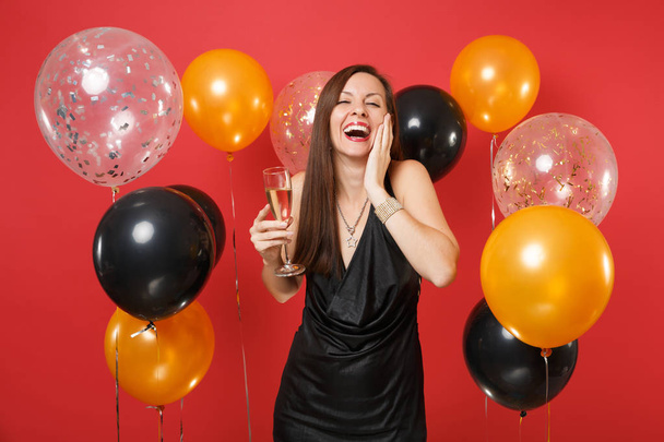 Γέλιο κορίτσι στο μαύρο φόρεμα γιορτάζει κρατώντας το ποτήρι σαμπάνιας, κρατώντας το χέρι στο πρόσωπό σε λαμπερό κόκκινο φόντο και αερόστατα. Ημέρα του Αγίου Βαλεντίνου, ευτυχισμένο το νέο έτος, γενέθλια mockup holiday κόμμα έννοια - Φωτογραφία, εικόνα