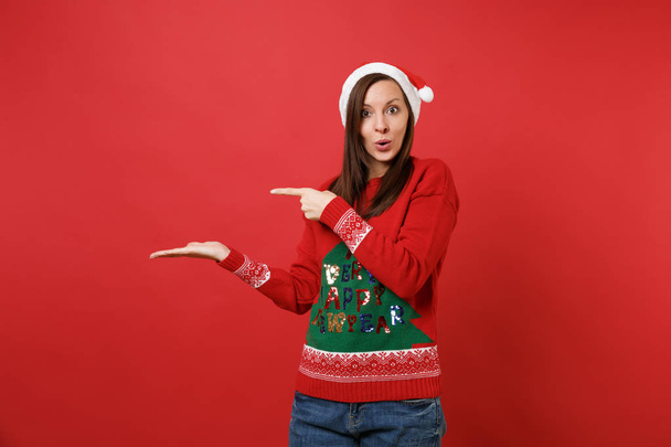 Amazed Santa girl in knitted sweater, Christmas hat looking surprised pointing hand, finger aside isolated on red background. Happy New Year 2019 celebration holiday party concept. Mock up copy space - Φωτογραφία, εικόνα
