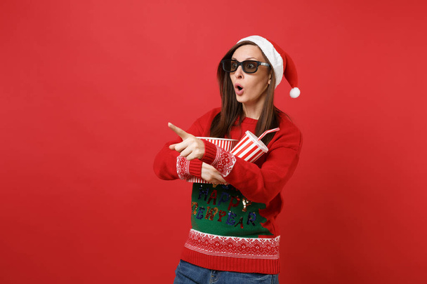 Shocked young Santa girl in 3d imax glasses watching movie film, hold popcorn, cup of soda pointing index finger isolated on red wall background. Happy New Year 2019 celebration holiday party concept - Foto, afbeelding