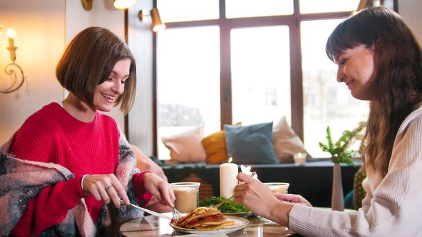 Két fiatal, mosolygós nő ül a modern kávézó és egy reggelizik. Palacsinta és a kávé - Fotó, kép