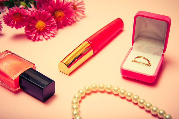 Golden ring in box, beads, nail polish, lipstick and flowers on a pink background. Women jewelry and accessories. Color toned. - Photo, image