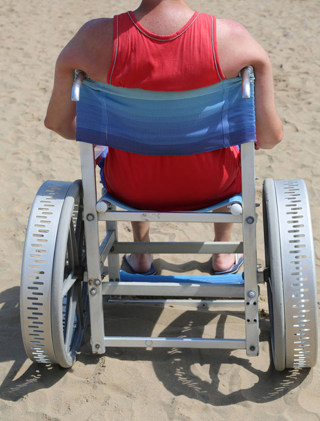 joven discapacitado con camiseta en la silla de ruedas en la playa de arena del pueblo turístico en el verano
 - Foto, imagen