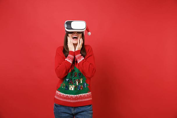 Удивительная девушка Санта в трикотажном свитере, рождественская шляпа, смотрящая в наушники, кладущая руки на лицо, изолированное на красном фоне. Концепция праздничной вечеринки в честь Нового 2019 года. Пространство для копирования
 - Фото, изображение
