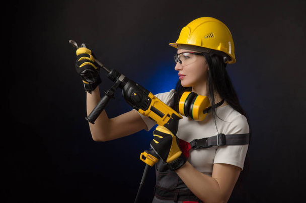 the brunette girl in special clothes and a worker in a helmet posing on a black background with a working tool ( perforator, drill, percussion hammer ) - Photo, Image
