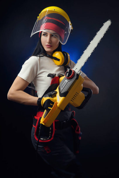 the brunette girl in special clothes and a worker in a helmet posing on a black background with a working tool ( power saw ) - Photo, Image