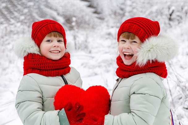 双子姉妹を笑いに身を包んだ明るい服が心から一緒に冬公園の背景を手に大きな赤いハートを持って笑っています。コンセプトは、聖バレンタインの日 - 写真・画像