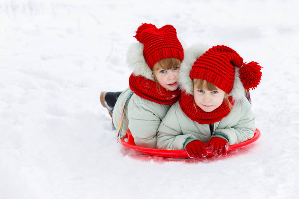 Смішні сестри близнюки лежать на пластиковій тарілці, насолоджуючись катанням на санчатах зі сніжного пагорба в зимовому парку. Концепція дитячої гри взимку
. - Фото, зображення