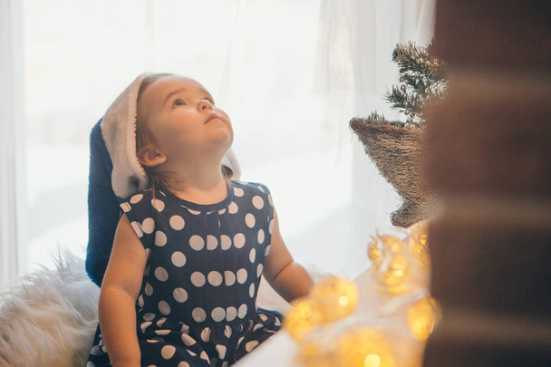 a little girl in a blue Christmas hat sitting near the battery and a table with a garland and a small Christmas tree - Фото, изображение