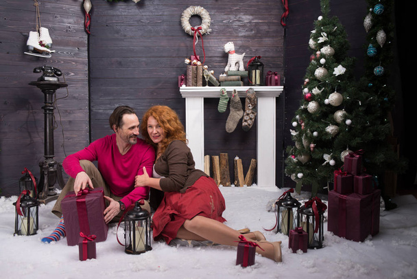 Longitud completa de encantadora pareja madura feliz sentada de cerca en una alfombra suave cerca del árbol de Navidad mientras está lista para desempacar regalos
 - Foto, Imagen
