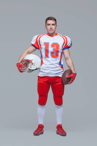 American football player holding rugby ball and helmet against grey background full length - Foto, Imagem