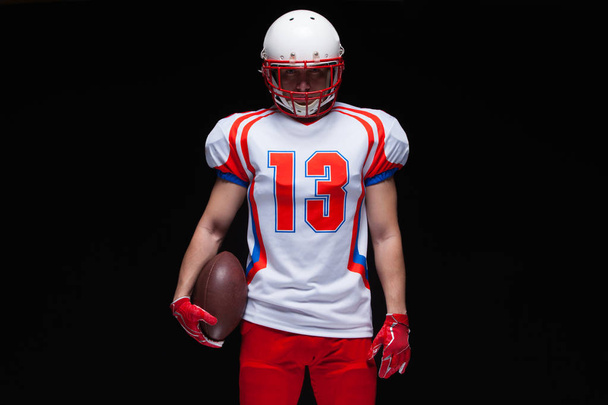 American football player wearing helmet posing with ball on black background - Valokuva, kuva