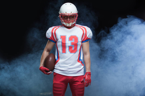 Front view portrait of american football player wearing helmet with ball against white smoke - Фото, изображение