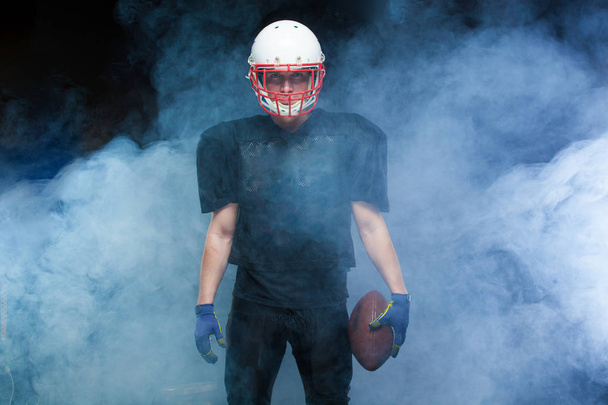 American football player in a black uniform, wearing helmet and holding ball against white smoke - Foto, imagen