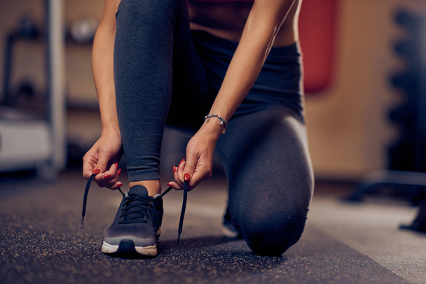 Primo piano della donna sportiva caucasica che lega il lacci delle scarpe mentre si inginocchia. Interni palestra, concetto di stile di vita sano
. - Foto, immagini