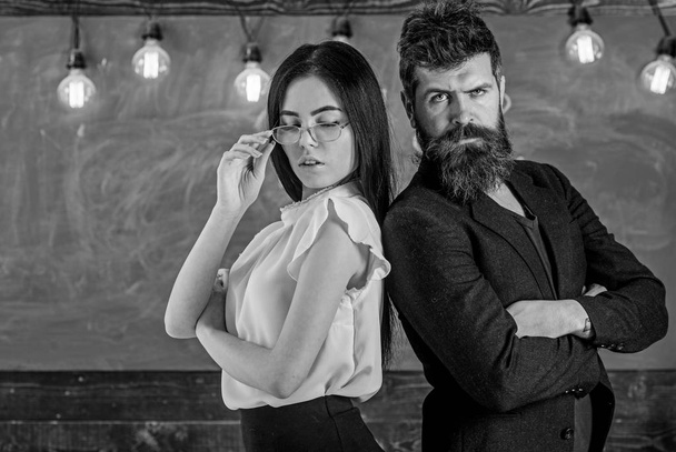 Man with beard and teacher in eyeglasses stand back to back, chalkboard on background. Teamwork concept. Teacher and schoolmaster look confident. Lady and hipster working together in school - Zdjęcie, obraz