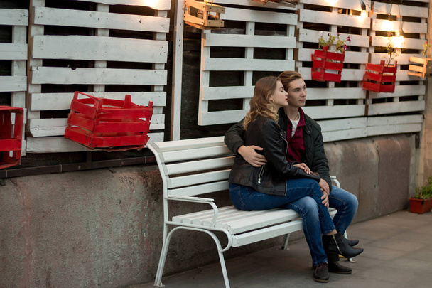 Abendtermin. Verliebte Paare sitzen auf einer weißen Bank vor dem Hintergrund weißer Bretter und Kästen unter den Blumen - Foto, Bild