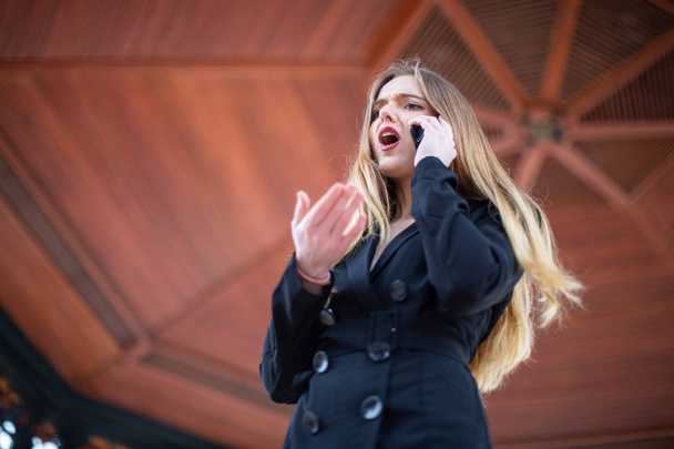 Mooie jonge vrouw in elegante outfit schreeuwen en gebaren terwijl ontslagen van werk telefonisch in park - Foto, afbeelding