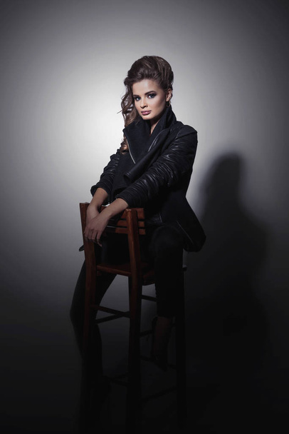 Стильная молодая модель в черной одежде, сидящая на стуле и позирующая на сером фоне
 - Фото, изображение