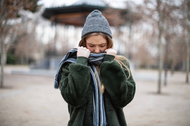 hübsche junge Frau in warmen Kleidern, die sich in Schal hüllt und wegschaut, während sie an einem wirklich kalten Tag vor dem verschwommenen Hintergrund des Herbstparks steht  - Foto, Bild