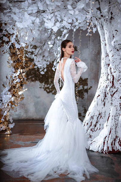 Fashion portrait of beautiful young woman in a gorgeous wedding dress, posing in a fantasy interior with white winter tree - Foto, Bild